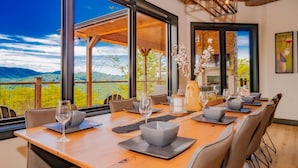 A dining area with a long table set for six, large windows offering a view of the countryside, and a modern interior design. - Manna Mountain Lodge ~  Indoor Pool, XBox Room, Gaming Garage, Views & More! (Sevierville)