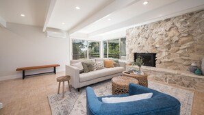 A modern living room with a stone fireplace, a sofa, a blue armchair, and a bench. - Mid-Century Retreat Overlooking the Pines & Pacific (Cambria)