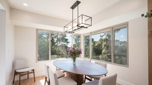 A dining area with a round table, six chairs, a vase with flowers, and large windows offering a view of trees and buildings. - Mid-Century Retreat Overlooking the Pines & Pacific (Cambria)