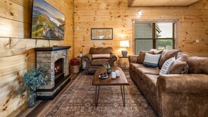 A cozy log cabin interior with a stone fireplace, leather furniture, and a large painting on the wall. - 3 Miles to Pigeon Forge Parkway! Private Pool 🏊‍♂️ Hot Tub 🐕 Dogs 🏔️ GSMNP Views 🌄 (Sevierville)