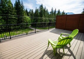 Una terraza de madera con dos sillas Adirondack verdes, una valla de madera y vistas a un bosque y montañas.