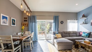 Una sala moderna con comedor, un sofá y una mesa de centro. Hay un ventanal con cortinas, un reloj en la pared y un suelo de madera con un diseño geométrico.