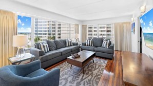 Ein modernes Wohnzimmer mit einem Ecksofa, einem Couchtisch und einem Fernseher. Große Fenster bieten Blick auf die Stadtlandschaft.