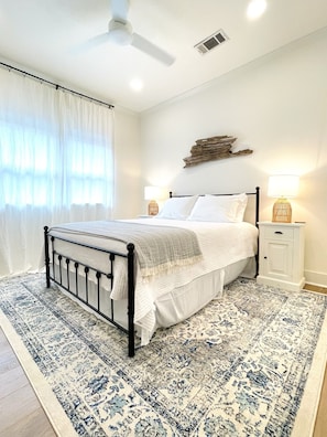 A bedroom with a bed, two bedside tables, a ceiling fan, a window with curtains, and a decorative wall piece.