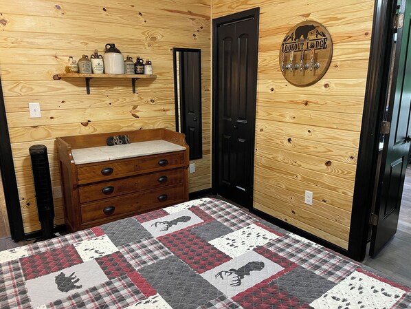 Quarto de madeira com cômoda de madeira, colcha acolchoada, prateleira com garrafas e porta com placa.