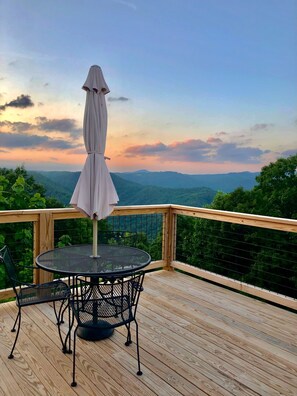 Um deck de madeira com uma mesa e cadeiras de metal redondas, um guarda-sol e vista para uma paisagem montanhosa.