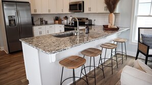 A kitchen with white cabinets, a stainless steel refrigerator, a granite countertop, a sink, a microwave, and a vase with dried plants. - Beautiful Beau Coast Home w/ Pool, Bikes, EV Charger & Dock Access (Beaufort)