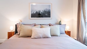 Une chambre à coucher avec un grand lit, deux tables de chevet avec des lampes, un tableau encadré au mur et une fenêtre avec des rideaux.