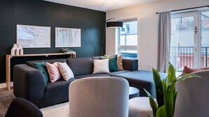 Un salon moderne avec un mur d’accent vert foncé, un canapé gris agrémenté de plusieurs coussins, une table d’appoint en bois et deux œuvres d’art encadrées.