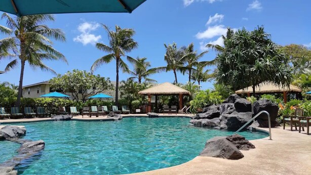 A swimming pool with clear water, surrounded by lounge chairs, palm trees, and thatched-roof structures. - Live Aloha! New furnished 2-bedroom Ko'olina (Kapolei)