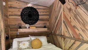 Un dormitorio con pared de listones de madera, cama con ropa blanca, un tapiz negro decorativo y un pequeño estante con un jarrón y una lámpara.