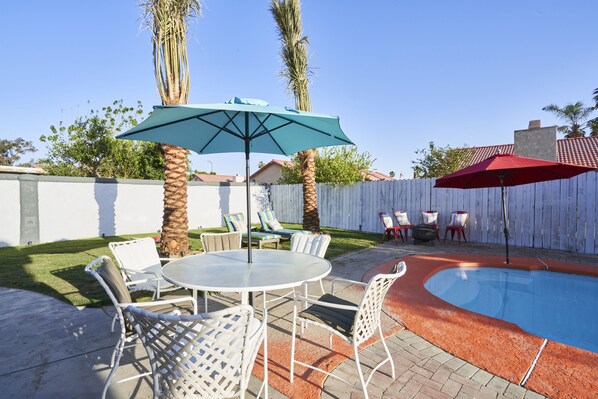 A backyard with a swimming pool, white outdoor furniture, and two umbrellas. - “The Place” Retro Oasis w/Pool, PacMan, & PingPong (Indio)