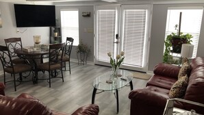 Un salon avec un canapé en cuir marron, une table basse en verre, une table à manger avec des chaises et un téléviseur à écran plat fixé au mur.