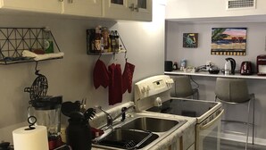 Une cuisine avec des meubles blancs, un four encastré, un évier et un plan de travail sur lequel se trouvent un mixeur et une bouilloire.