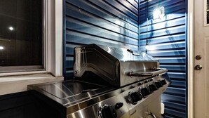 Un piano cottura a gas in acciaio inossidabile con quattro fuochi e una piastra, situato in una cucina dalle pareti azzurre orizzontali e con una finestra a sinistra.