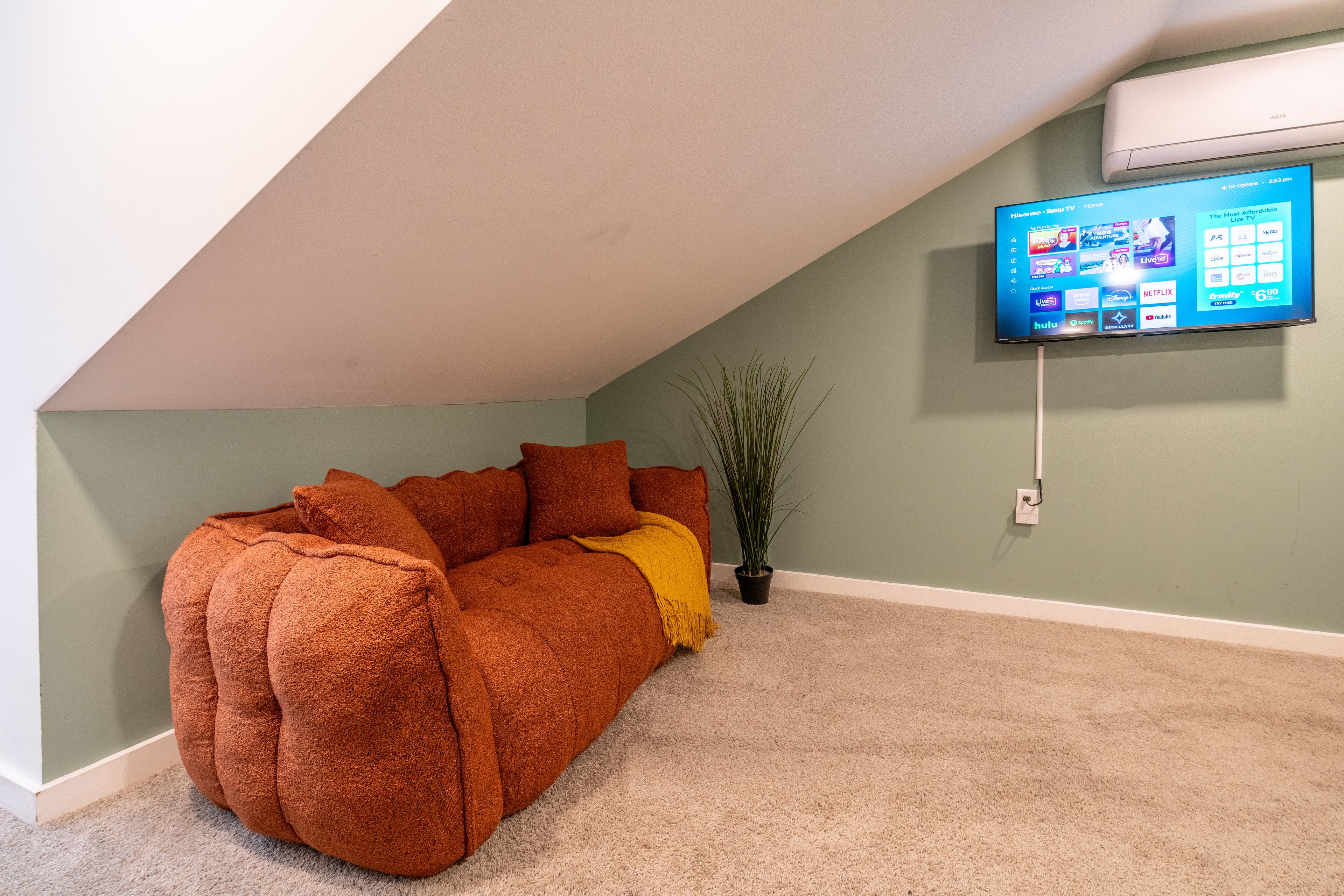Une pièce chaleureuse avec un plafond en pente, un canapé, une plante et un téléviseur fixé au mur, qui diffuse différents services de streaming.