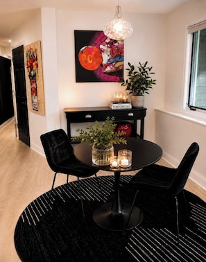 Un comedor con una mesa redonda, sillas negras, un tapete negro, una planta en maceta, una obra de arte colgada en la pared y una lámpara colgante.
