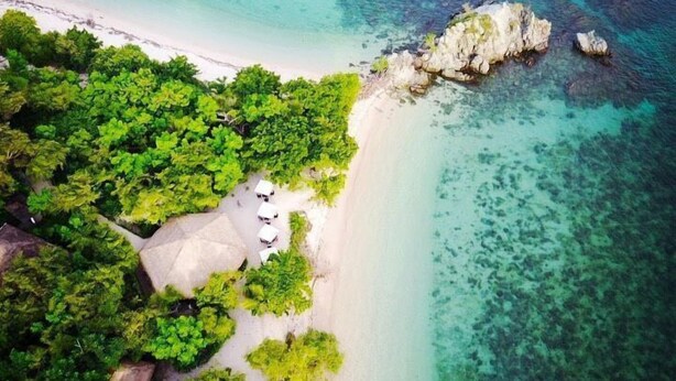 Eine Vogelperspektive zeigt einen Strand mit klarem türkisblauem Wasser, üppiger grüner Vegetation und einigen Gebäuden mit Reetdächern.