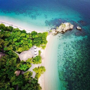 An aerial view of a beach with clear turquoise waters, lush green vegetation, and a few structures with thatched roofs. - Angel Island Eco Resort (Bidadari Island)