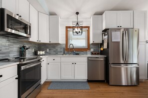 A modern kitchen with stainless steel appliances, white cabinetry, a wooden floor, and a window with blinds. - Just Wright: Heated Pool, Soundside, close to beach & sound! (Kitty Hawk)