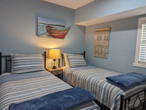 A bedroom with two beds, a nightstand with a lamp, a wall painting of a boat, and a macrame wall hanging. - Just Wright: Heated Pool, Soundside, close to beach & sound! (Kitty Hawk)