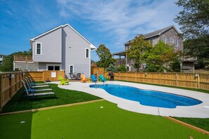 A backyard with a swimming pool, artificial grass, and a wooden deck with chairs. - Just Wright: Heated Pool, Soundside, close to beach & sound! (Kitty Hawk)