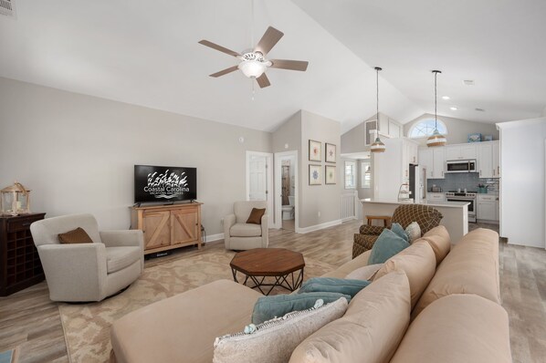 A spacious living room with a sofa, armchairs, a coffee table, and a television. There is a ceiling fan, a kitchen area in the background, and a large window.