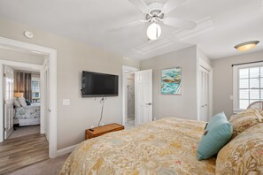 A bedroom with a bed, a ceiling fan, a mounted TV, a painting, and a window with blinds.