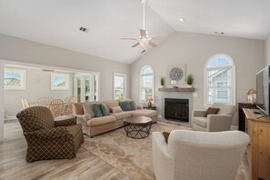 A spacious living room with a fireplace, a ceiling fan, and large windows.