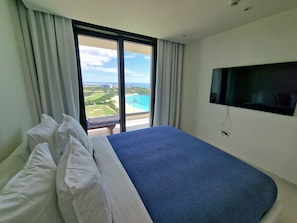 A hotel room with a large bed, a TV mounted on the wall, and a balcony with a view of the beach and ocean. - A-1602 Modern oceanfront condo (Cupecoy)
