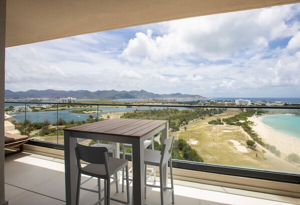 A balcony with a table and chairs overlooking a beach, mountains, and a bridge.