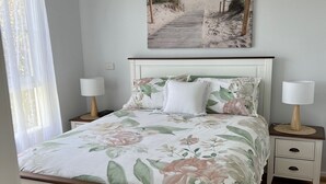 Ein Schlafzimmer mit einem Bett, zwei Nachttischen, einem Fenster mit transparenten Vorhängen und einem Gemälde mit einer Strandansicht an der Wand.