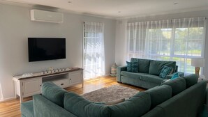 Ein modernes Wohnzimmer mit Flachbildfernseher, weißem Konsolentisch, einer grünen Sofagarnitur und großen Fenstern mit transparenten Vorhängen.