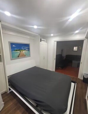 Une chambre à coucher avec un lit, un tableau représentant une plage, une armoire et une porte donnant sur une autre pièce.