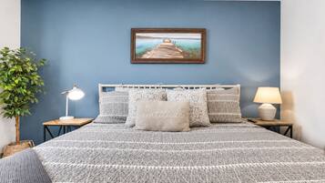 A bedroom with a bed, a nightstand, a lamp, a framed picture on the wall, and a plant.
