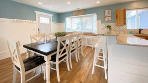 A dining area with a dark wooden table and white chairs, a kitchen with white cabinets and a window, and a sign reading "Beach House".