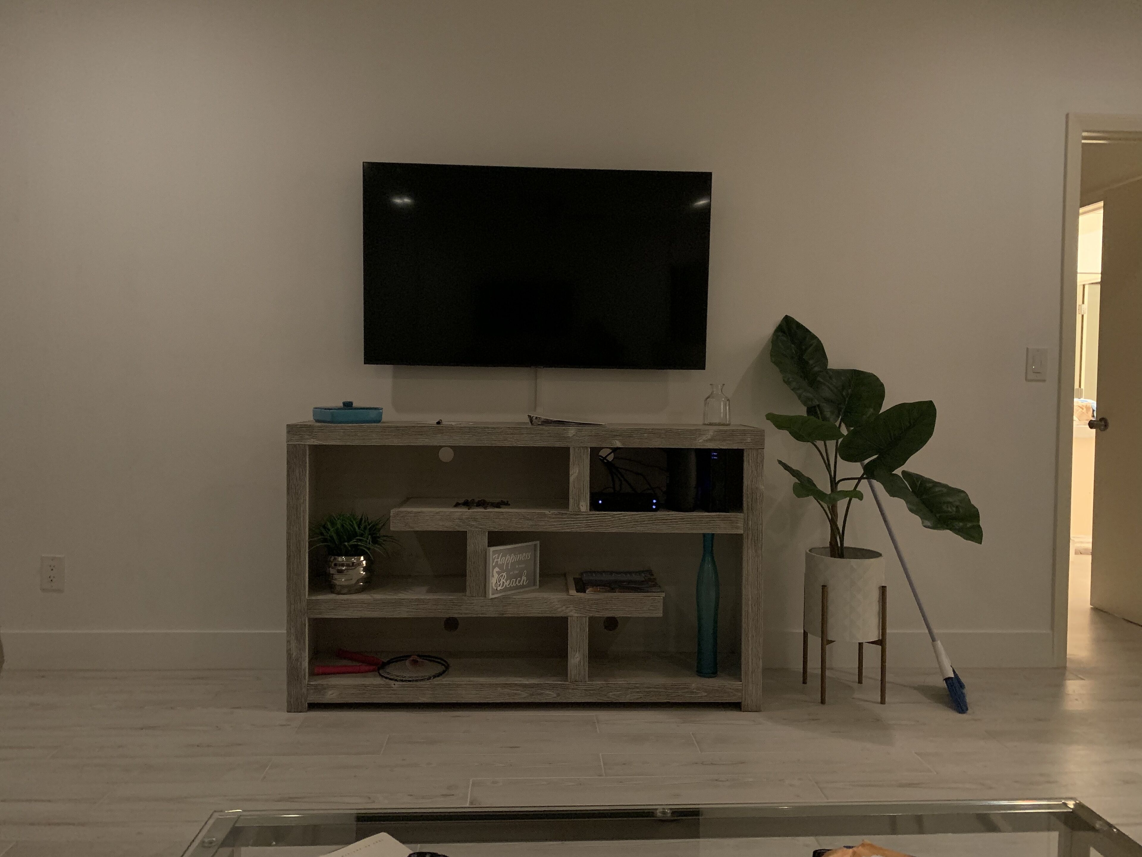 A living room with a flat-screen TV mounted on the wall, a wooden entertainment unit with shelves, a potted plant, and a mop leaning against the wall.