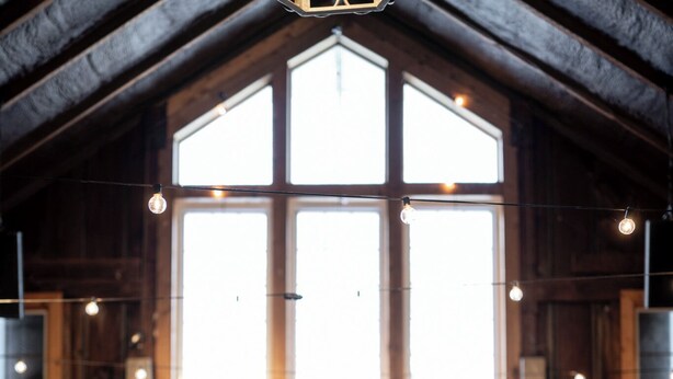 A rustic interior with a large wooden ceiling, a cozy seating area with a dark sofa and armchair, and a large window with string lights.