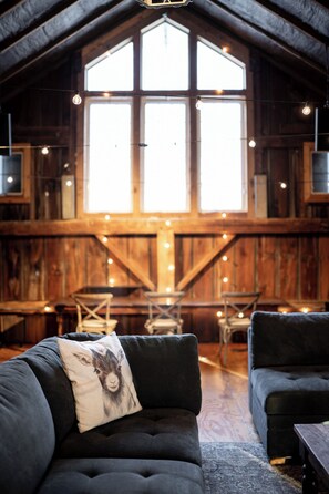 Un interior rústico con un amplio techo de madera, un rincón de descanso acogedor con un sofá y un sillón oscuros, y un ventanal con luces colgantes.