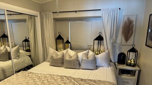 Un dormitorio con una cama grande, varios almohadones, una mesita de noche con un reloj y velas, y un espejo.