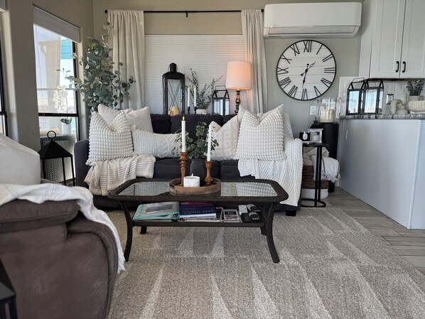 Un salon chaleureux avec une grande horloge murale, une table basse avec des livres et des télécommandes, un canapé noir et un fauteuil blanc.