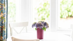 A white table with a purple vase containing purple flowers, set against a window with white curtains.