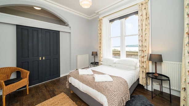 A bedroom with a large bed, a chair, two bedside tables, a window with curtains, and a view of the sea.