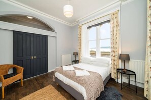 A bedroom with a large bed, a chair, two bedside tables, a window with curtains, and a view of the sea. - The Gate Lodge - Modern & Period, Combined (Broughty Ferry, Dundee)