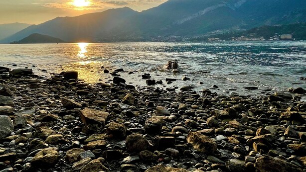 A rocky shoreline with a mountain backdrop and a setting sun.