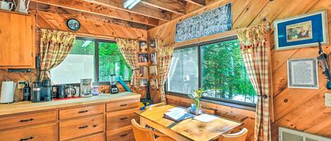 Ein Holzblockbau-Innenraum mit Esstisch, Stühlen, einer Küchenzeile inklusive Geräte und einem großen Fenster mit Vorhängen.
