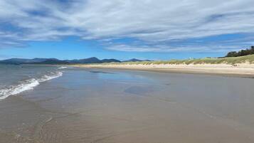 A wide sandy beach with gentle waves, a clear sky, and distant mountains.