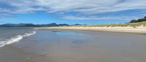Une vaste plage de sable fin, avec des vagues douces, un ciel dégagé et des montagnes à l’horizon.