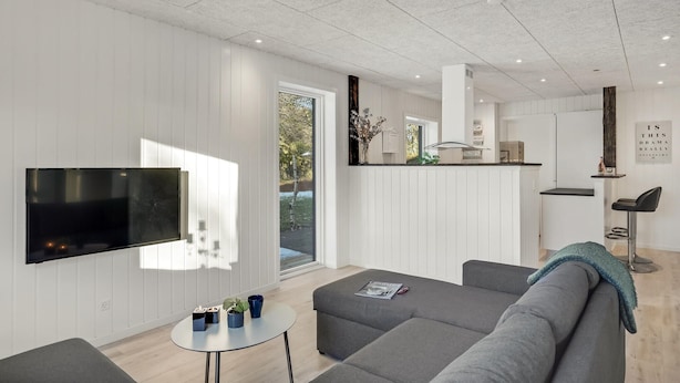 A modern living room with a flat-screen TV, grey sofas, a small round table, and a kitchen area with a countertop and cabinets. - 3 bedroom amazing home in Storvorde (Storvorde)