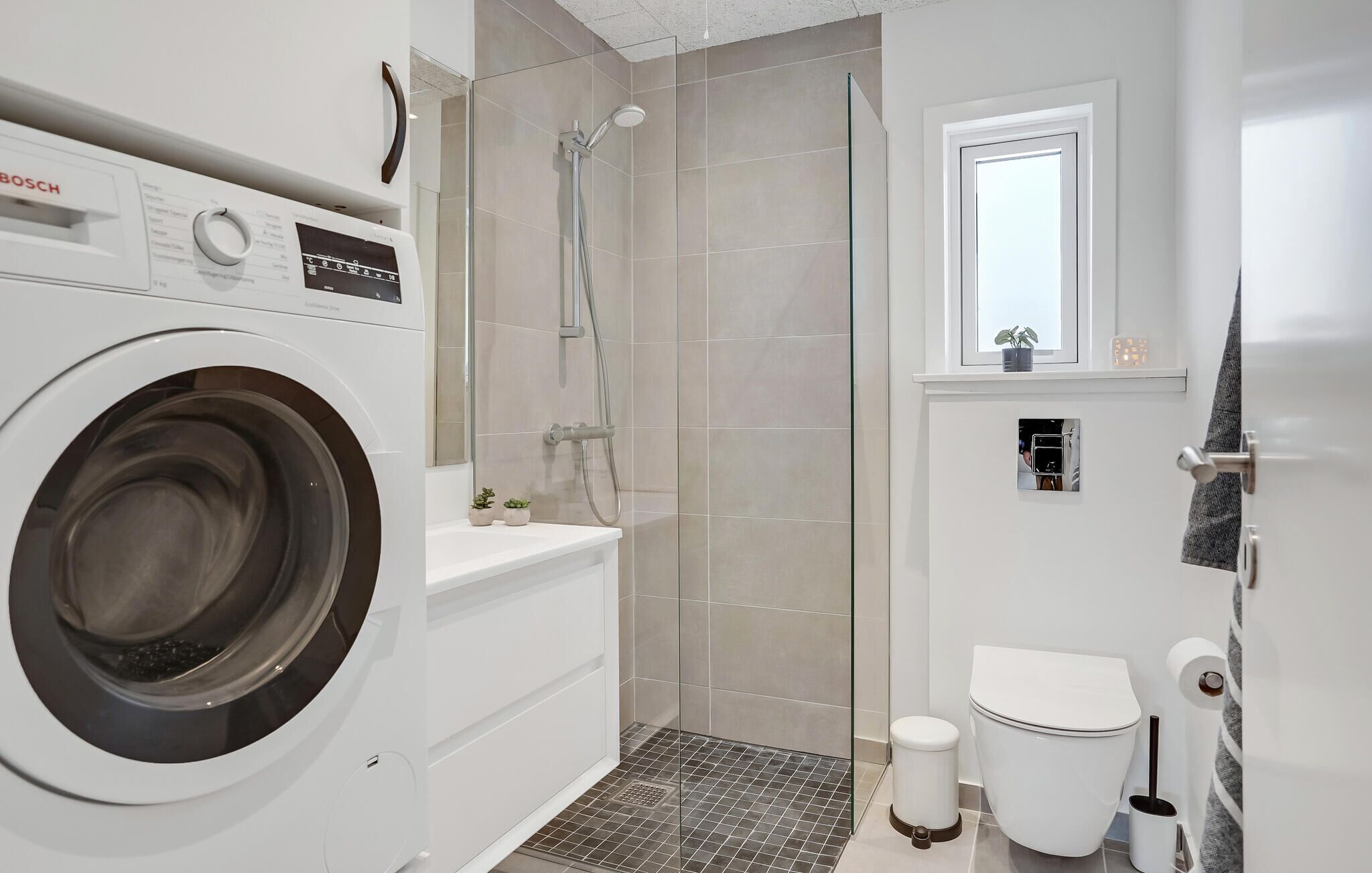 A modern bathroom with a washing machine, a glass shower enclosure, a toilet, and a small window.